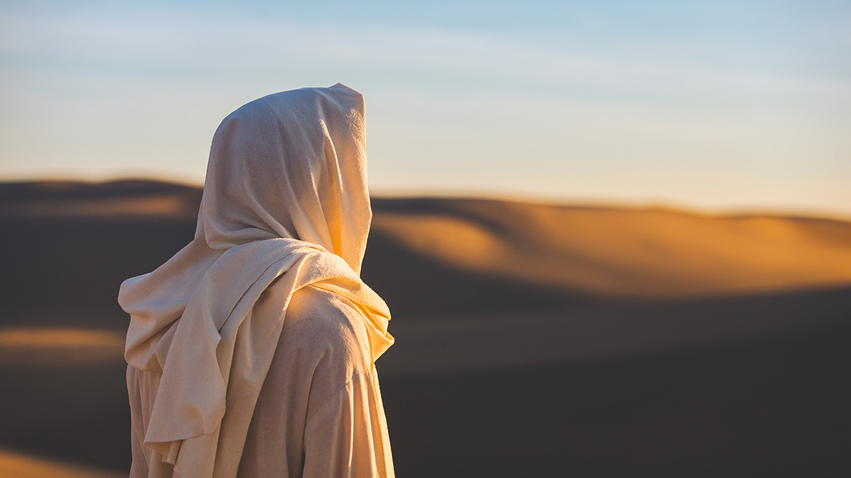 Jesus Christ looks out at a setting sun in the sand dunes.